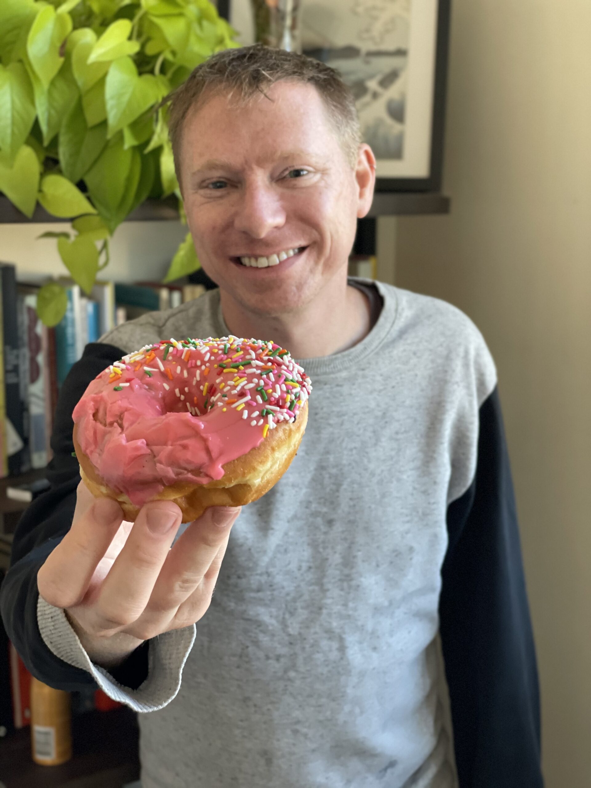 photo of smiling man in gray sweater holding a pink sprinkle donut in the foreground